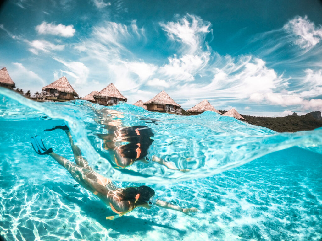 A snorkeler swimming in crystal-clear turquoise waters with overwater bungalows and a scenic mountain in the background at Bora Bora.